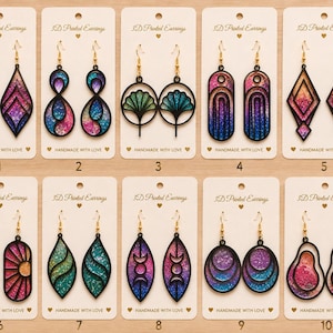 Puede incluir: Diez pares de pendientes impresos en 3D presentados en tarjetas blancas. Cada par tiene marcos negros con centros de colores degradados y brillantes en varias formas geométricas, florales y abstractas, colgados de ganchos de metal dorado.