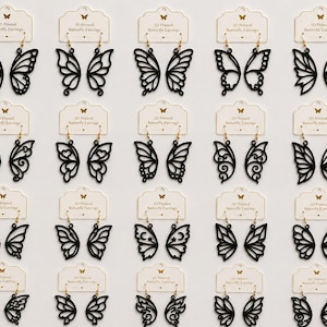 Può includere: Una collezione di venti paia di orecchini a forma di ali di farfalla stampati in 3D in nero, montati su cartoncini bianchi con l'etichetta 3D Printed Butterfly Earrings. Gli orecchini presentano vari motivi traforati e ganci color oro.