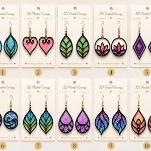 Puede incluir: Diez pares de pendientes impresos en 3D presentados en tarjetas blancas. Cada par cuenta con marcos negros con inserciones translúcidas, coloridas y brillantes en formas de hojas, corazones, flores de loto, diamantes y ondas circulares.