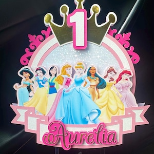 Pode incluir: Topper de bolo com tema de princesas, com o número 1 e o nome "Aurelia". O design inclui imagens de princesas da Disney, em um fundo rosa e dourado. Uma coroa está no topo.