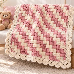 Puede incluir: Una manta de ganchillo con un patrón de tejido de cesta en colores alternos de hilo rosa empolvado y crema. La manta rectangular está rematada con un borde decorativo festoneado de color crema que recorre los cuatro lados del tejido.