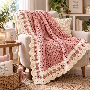 May include: A crochet blanket featuring a dusty rose center with a textured stitch pattern, bordered by a wide cream-colored trim decorated with small rose-colored flower motifs and a scalloped edge, draped over a light-colored upholstered chair.
