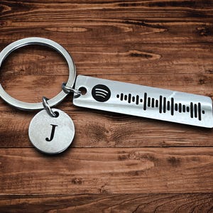 May include: A silver keychain with a round tag featuring the letter "J" and a rectangular tag with a Spotify code. The keychain is on a wooden surface.