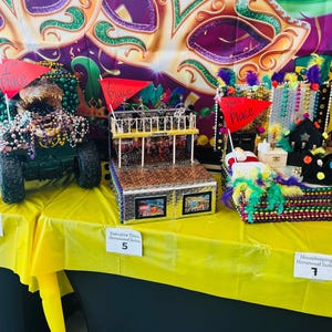 Könnte beinhalten: Drei dekorative Festwagen auf einem gelben Tisch, verziert mit Perlen, Flaggen und verschiedenen Verzierungen. Die Festwagen sind mit Platzierungsnummern und Teamnamen versehen und präsentieren eine festliche und kreative Ausstellung.