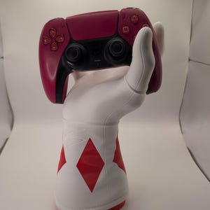 May include: A magenta and black wireless video game controller held by a white, sculpted hand stand. The base of the stand is white with red diamond-shaped accents, featuring a smooth, matte finish and a structured, glove-like design.