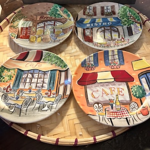Könnte beinhalten: Ein Set aus vier runden Keramiktellern mit farbenfrohen Illustrationen von Ladenfronten im europäischen Stil. Jeder Teller zeigt eine andere Szene mit Markisen, Tischen, Stühlen und Schriftzügen wie Bread, Bistro und Cafe in leuchtenden Farben.