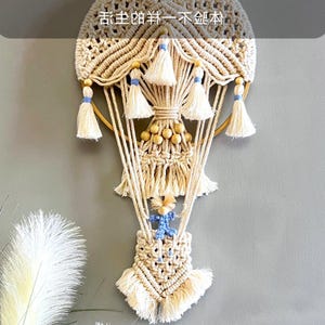 Könnte beinhalten: Handgefertigter Makramee-Wandbehang in Beige und Creme, mit einem Heißluftballon-Design. Das Stück enthält Quasten, Holzperlen und eine kleine blaue Figur. Der obere Bereich hat chinesischen Text.