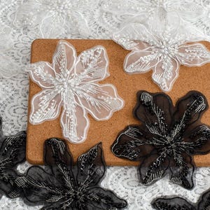 May include: Close-up of floral appliques in white and black. The white appliques feature intricate beadwork and a sheer outline. The black appliques have a sequined appearance. The appliques are arranged on a corkboard and lace fabric.