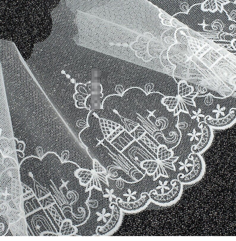 Peut inclure: Un tissu en tulle blanc avec une bordure en dentelle brod&eacute;e complexe. Le motif se compose de ch&acirc;teaux r&eacute;p&eacute;t&eacute;s entour&eacute;s de formes nuageuses, de petites &eacute;toiles et d'accents en forme de papillon le long d'un bord festonn&eacute; avec des d&eacute;tails floraux.