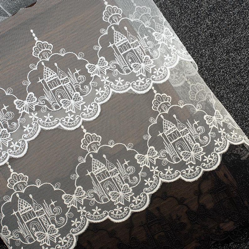 Peut inclure: Un tissu en dentelle blanche pr&eacute;sentant un motif r&eacute;p&eacute;titif de ch&acirc;teaux d&eacute;taill&eacute;s entour&eacute;s de bordures festonn&eacute;es. Le design complexe inclut de petites formes d'&eacute;toiles et des accents floraux, la dentelle blanche &eacute;tant superpos&eacute;e sur un fond sombre.