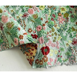 May include: A piece of fabric featuring a dense, multi-colored floral pattern with embroidered-style textures. The design includes various shades of green, pink, red, and yellow flowers and foliage on a light background, with a folded corner.