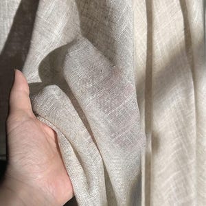 May include: Close-up of a light beige linen fabric. The fabric has a textured weave and is draped, with shadows creating depth. The material appears soft and lightweight, suitable for clothing or home decor.
