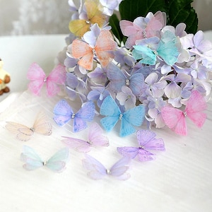 May include: Assortment of pastel colored butterfly embellishments with rhinestones. The butterflies are made of sheer fabric and are arranged on a white surface.