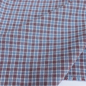May include: Close-up of a fabric with a blue, white, and burgundy plaid pattern. The fabric is folded, showing the texture and weave of the material. The edges are frayed, indicating a cut piece of fabric.