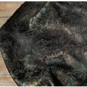Può includere: Primo piano di un tessuto nero con un dettagliato motivo floreale in pizzo. Il tessuto ha una sottile lucentezza con sfumature oro e verde, creando un aspetto strutturato e lussuoso. Il bordo del tessuto è visibile.