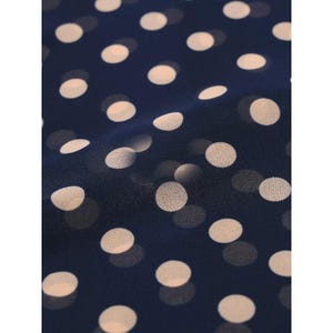 May include: A dark navy blue fabric featuring a repeating pattern of soft, light beige polka dots. The material has a slightly sheer, lightweight texture with gentle folds that create subtle shadows and depth across the dotted surface.