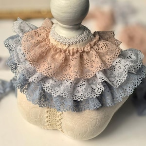 Könnte beinhalten: Ein mehrlagiger Spitzenkragen mit drei Reihen in den Farben Rosa, Blau und Weiß. Der Kragen ist aus zarter Spitze gefertigt und hat einen gekräuselten Rand.