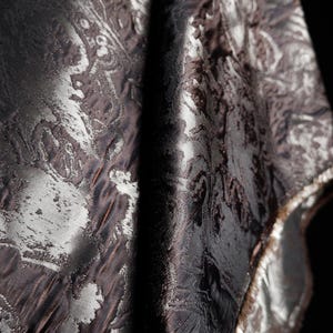 Może przedstawiać: Zbliżenie ciemnobrązowej i srebrnej tkaniny we wzory. Tkanina ma teksturowaną powierzchnię z wirującym, kwiatowym wzorem. Krawędzie są obszyte srebrną lamówką. Tkanina jest udrapowana, tworząc cienie i światła.