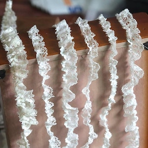 Puede incluir: Primer plano de varios hilos de encaje blanco con un borde con volantes. El encaje es delicado y tiene un patrón floral. El adorno se muestra sobre una superficie de madera.