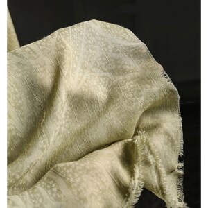 May include: Close-up of a light green fabric with a subtle, repeating pattern. The fabric has a slightly textured appearance and a fringed edge. The material is draped, showing folds and shadows.