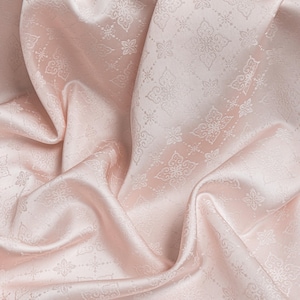 May include: A pale pink fabric featuring a repeating damask-style pattern of intricate floral and geometric motifs. The material has a smooth, lustrous finish with soft folds and drapes that create subtle highlights and shadows across the surface.