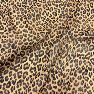 Puede incluir: Primer plano de una tela con estampado de leopardo. La tela tiene un fondo beige con manchas marrones oscuras y negras. La tela está drapeada, mostrando pliegues y textura.