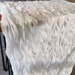 Könnte beinhalten: Ein weißer Tischläufer mit einer stark texturierten, zerknitterten und gerafften Stoffoberfläche. Das Material hat eine transparente, halb-durchscheinende Qualität mit vertikalen Wellen und Falten, die ein geschichtetes, voluminöses Erscheinungsbild erzeugen.
