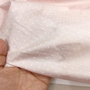 May include: Close-up of a pale pink fabric with a pattern of small white polka dots. The fabric is gathered and folded, creating shadows and texture. The fabric is being held by a hand.