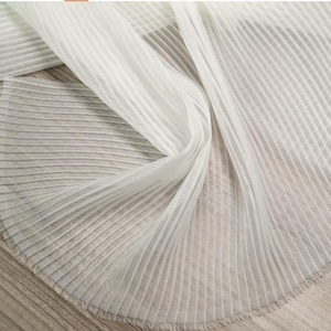 May include: A sheer white fabric featuring a fine, vertical pleated texture. The material is lightweight and semi-transparent, with consistent parallel folds running throughout the length of the textile, creating a structured yet flowing appearance.