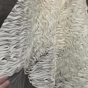 Puede incluir: Primer plano de tela blanca con textura ondulada y en capas. La tela parece ser un material delicado, posiblemente para un vestido u otra prenda. El diseño presenta un patrón repetitivo de elementos con volantes en forma de cinta.
