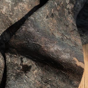 Könnte beinhalten: Nahaufnahme eines strukturierten Stoffes mit dunklem, gesprenkeltem Aussehen. Das Material hat ein komplexes Muster mit Schattierungen von Schwarz, Braun und Goldtönen, wodurch eine robuste, gealterte Ästhetik entsteht. Der Stoff scheint gefaltet zu sein und zeigt seine geschichtete Konstruktion.