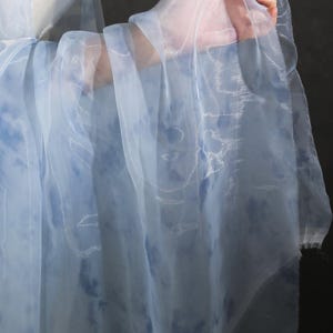 Puede incluir: Tejido transparente azul claro y blanco con efecto acuarela. La tela se sostiene, mostrando su delicada textura y su caída suave y fluida. La tela tiene un patrón sutil.