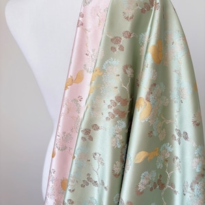 Puede incluir: Dos piezas de tela drapeadas sobre un maniquí. Una es rosa pálido, la otra verde suave. Ambas tienen un patrón repetido de ardillas, ramas de pino y flores en oro, marrón y azul.