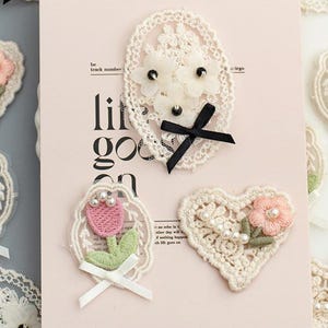May include: A set of five delicate lace appliques with floral designs and pearl accents. The appliques are in shades of white and pink and feature a variety of floral motifs, including roses, tulips, and daisies. The appliques are perfect for adding a touch of elegance to any craft project.