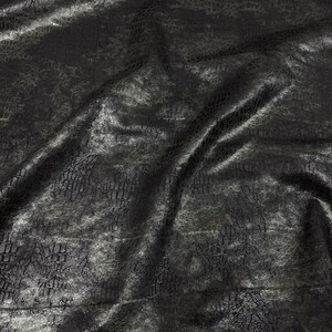 May include: A close-up of a black fabric with a textured, reptile-like pattern. The material has a shiny, reflective quality, with subtle variations in tone and highlights. The fabric is draped, creating folds and shadows.