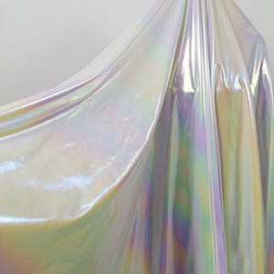 Può includere: Tessuto iridescente con finitura perlata e brillante. Il materiale riflette uno spettro di colori, tra cui sfumature di rosa, verde e giallo, creando un effetto dinamico e accattivante. Il tessuto appare liscio e fluido, con morbide pieghe.