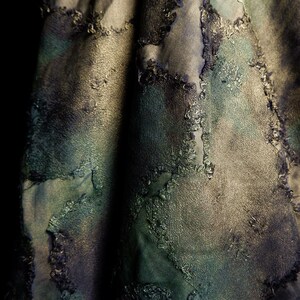 Op de afbeelding: Close-up van een textiel met een patchwork-ontwerp. De stof heeft een mix van donkergroene, blauwgroene en grijze tinten. De patches zijn omlijnd met een getrimde, gerafelde rand, waardoor een gelaagd effect ontstaat. De algehele esthetiek is artistiek en getextureerd.