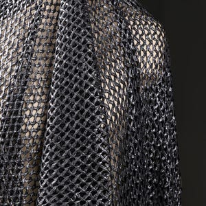 May include: Close-up of black, open-weave fabric with a metallic sheen. The fabric is draped, revealing a glimpse of a neutral-toned background. The texture is intricate, with a net-like pattern. This material could be used for apparel or decorative purposes.