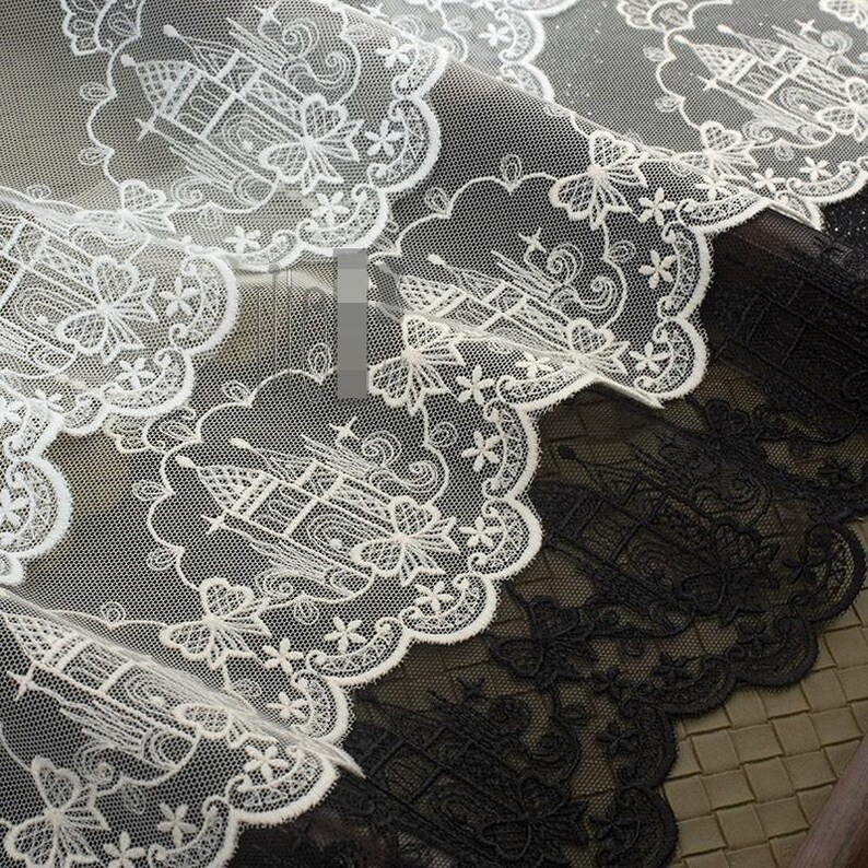 Peut inclure: Tissu en dentelle blanche pr&eacute;sentant un motif r&eacute;p&eacute;titif de ch&acirc;teaux brod&eacute;s &agrave; l'int&eacute;rieur de bordures festonn&eacute;es. Le design complexe comprend des accents floraux et un fond en maille fine. La dentelle est superpos&eacute;e sur un tissu noir assorti.