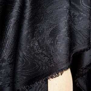 Puede incluir: Primer plano de una tela negra con un patrón texturizado y arremolinado. La tela tiene brillo y un borde deshilachado. El diseño se asemeja al agua o a un paisaje natural. Ideal para ropa o decoración del hogar.