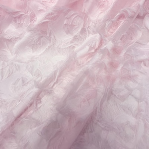 Puede incluir: Primer plano de una tela suave y fluida en tonos rosados. El material tiene una superficie delicada y texturizada con detalles plumosos y con flecos. La luz atrapa la tela, creando reflejos y sombras.