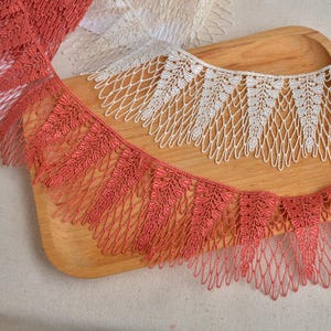 Puede incluir: Primer plano de dos adornos de encaje decorativos. Uno es de color coral, el otro es blanco. Ambos adornos presentan un diseño en forma de hoja a lo largo del borde superior y un patrón similar a una red con flecos. Los adornos están sobre una bandeja de madera.