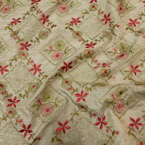 May include: Cream-coloured fabric with an embroidered floral pattern. The design features red and green flowers, along with gold-coloured diamond shapes. The fabric has a subtle sheen, giving it a luxurious appearance. The embroidery is detailed and intricate.