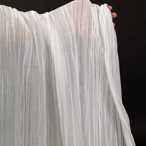 Pode incluir: Um tecido branco transparente com uma textura plissada vertical distinta. O material é leve e semitransparente, exibindo uma série de pregas finas e paralelas que vão da borda superior à inferior, criando um padrão linear consistente.