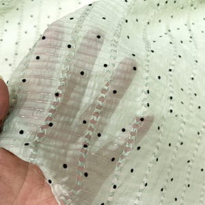 May include: A sheer, light green fabric with a dotted pattern and vertical striped accents. The fabric is held up, allowing light to pass through, revealing the texture and design. The dots are black and evenly spaced.