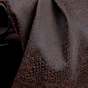 May include: Close-up of a dark brown fabric with a textured, mottled pattern. The fabric is draped, revealing folds and shadows. The material appears to be thick and possibly suitable for upholstery or drapery.