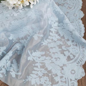 Puede incluir: Un camino de mesa de tela azul claro transparente con intrincados bordados florales y un borde de encaje festoneado. El material delicado muestra un patrón botánico repetitivo con hojas y flores detalladas en el borde.