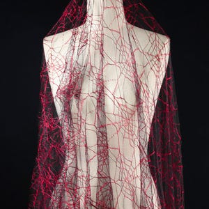 Puede incluir: Tela negra y roja con diseño de telaraña drapeada sobre un maniquí. La tela tiene una estructura delicada, similar a una red, con hilo rojo que crea el diseño de la telaraña. El número "36" es visible en la tela. El fondo es negro.