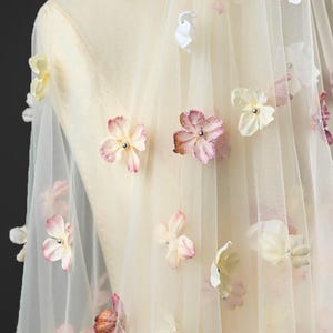 May include: Ivory tulle fabric adorned with delicate, three-dimensional flowers in shades of white, pale yellow, and pink. The floral embellishments are scattered across the sheer material, creating a romantic and ethereal effect. Ideal for bridal or special occasion wear.