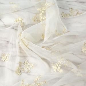 May include: A sheer, off-white fabric featuring delicate, scattered gold floral embroidery. The material is draped in soft folds, highlighting the translucent quality of the textile and the intricate, raised texture of the metallic gold leaf patterns.